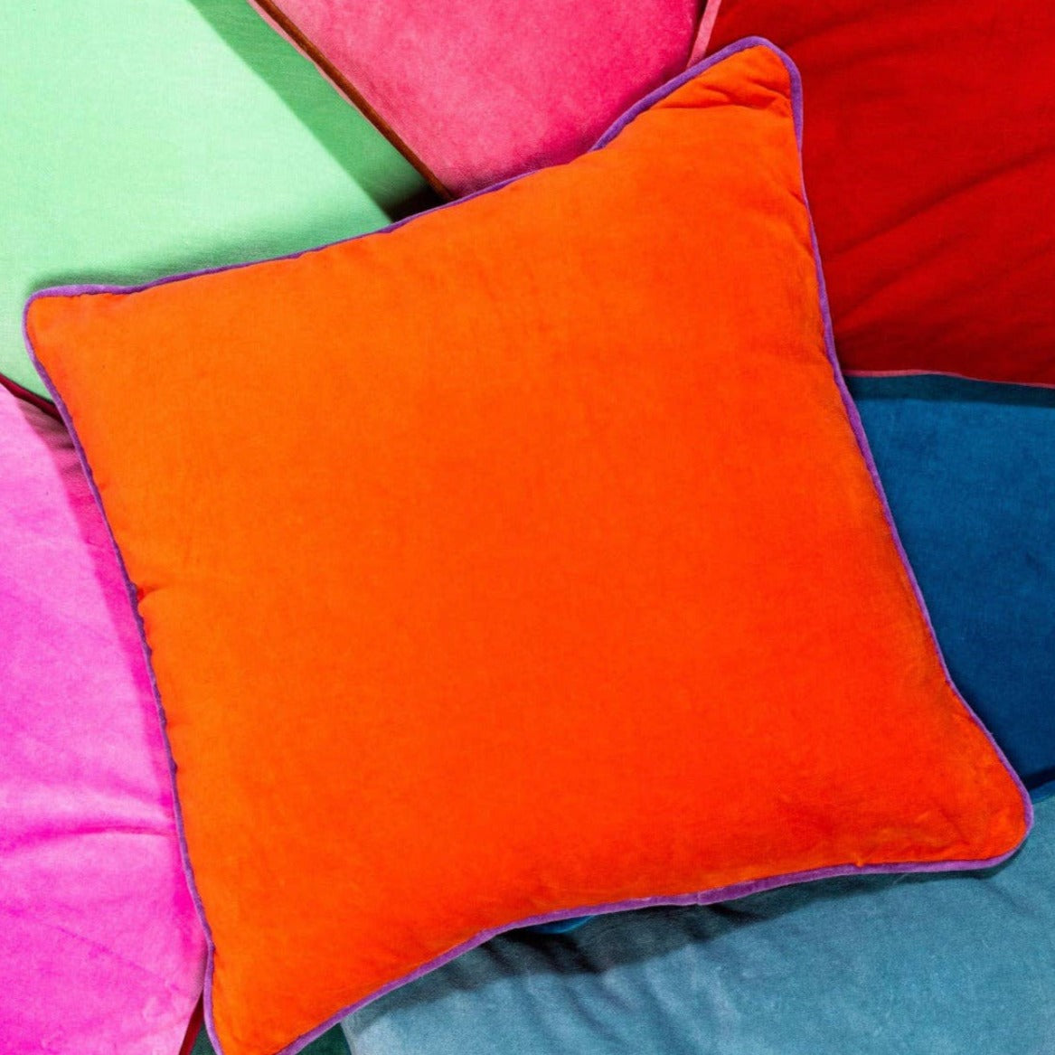 A bright orange velvet square pillow with a royal purple trim on the edges